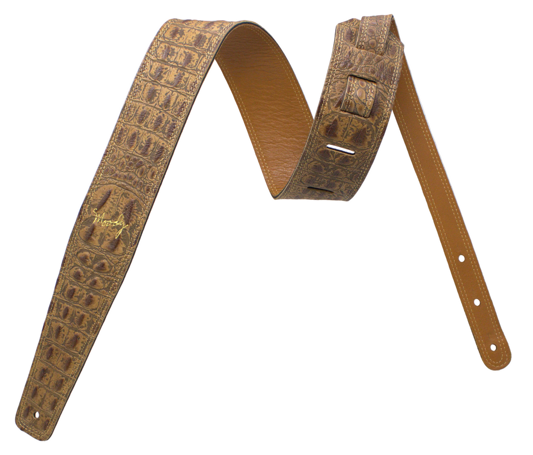 A tan leather guitar strap featuring an embossed crocodile pattern on one side and smooth leather on the other, with a gold-toned metal logo plate.